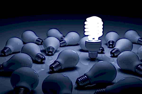 Compact Fluorescent Light Bulbs standing and shining in a dark room full of unlit, fallen conventional light bulbs