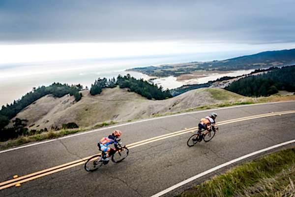Two bikers riding on a road near the ocean.