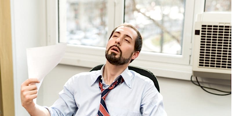 Hot office worker tries to fan himself with paper. Broken air conditioning unit behind him.