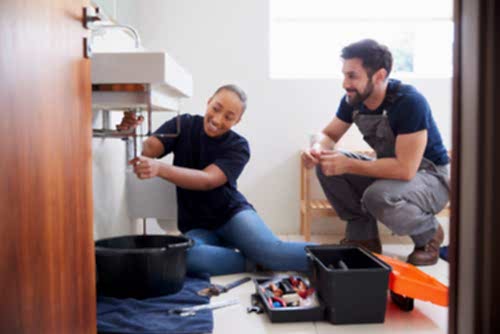 A plumber's apprentice works on the pipes beneath a sink while a licensed plumber supervises.