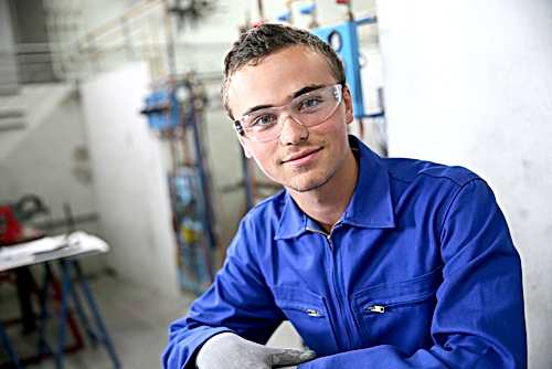 A young man in blue coveralls and safety glasses smiles during his plumbing apprenticeship.